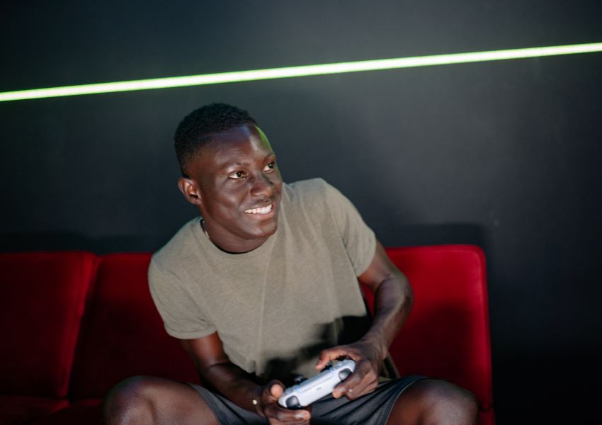 Smiling man sitting on a red couch holding a game controller in a dark room with neon lighting. Lifestyle scene representing online data gaming, fast internet, and entertainment connectivity.